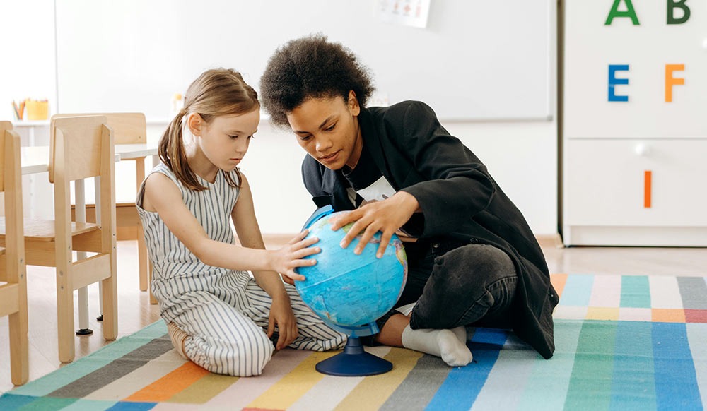An image of a female school counselor in a black jacket teaching a student how to read a globe. 
