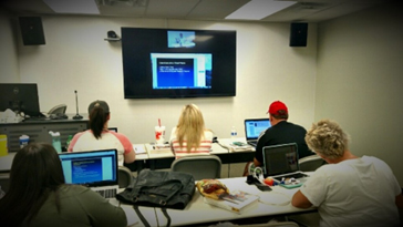 four students sitting in a classroom watching a remote lecture on a wall mounted screen 